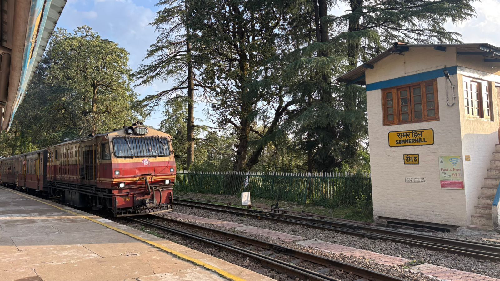 A red and cream narrow-gauge train standing at Summer Hill station on the Kalka-Shimla Railway, under tall deodar trees